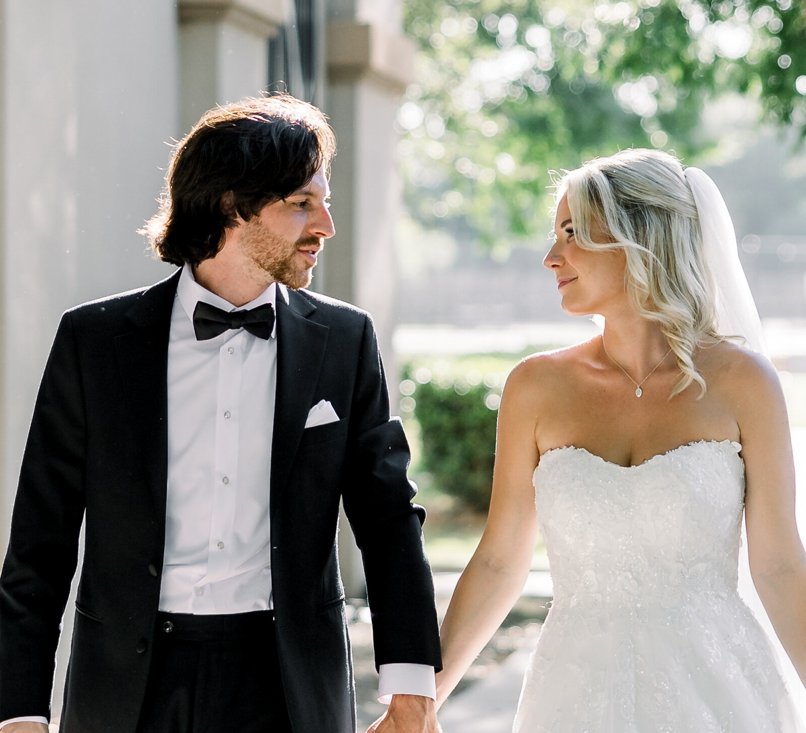 Bride and groom walking hand in hand outdoors, looking at each other lovingly in soft natural sunlight