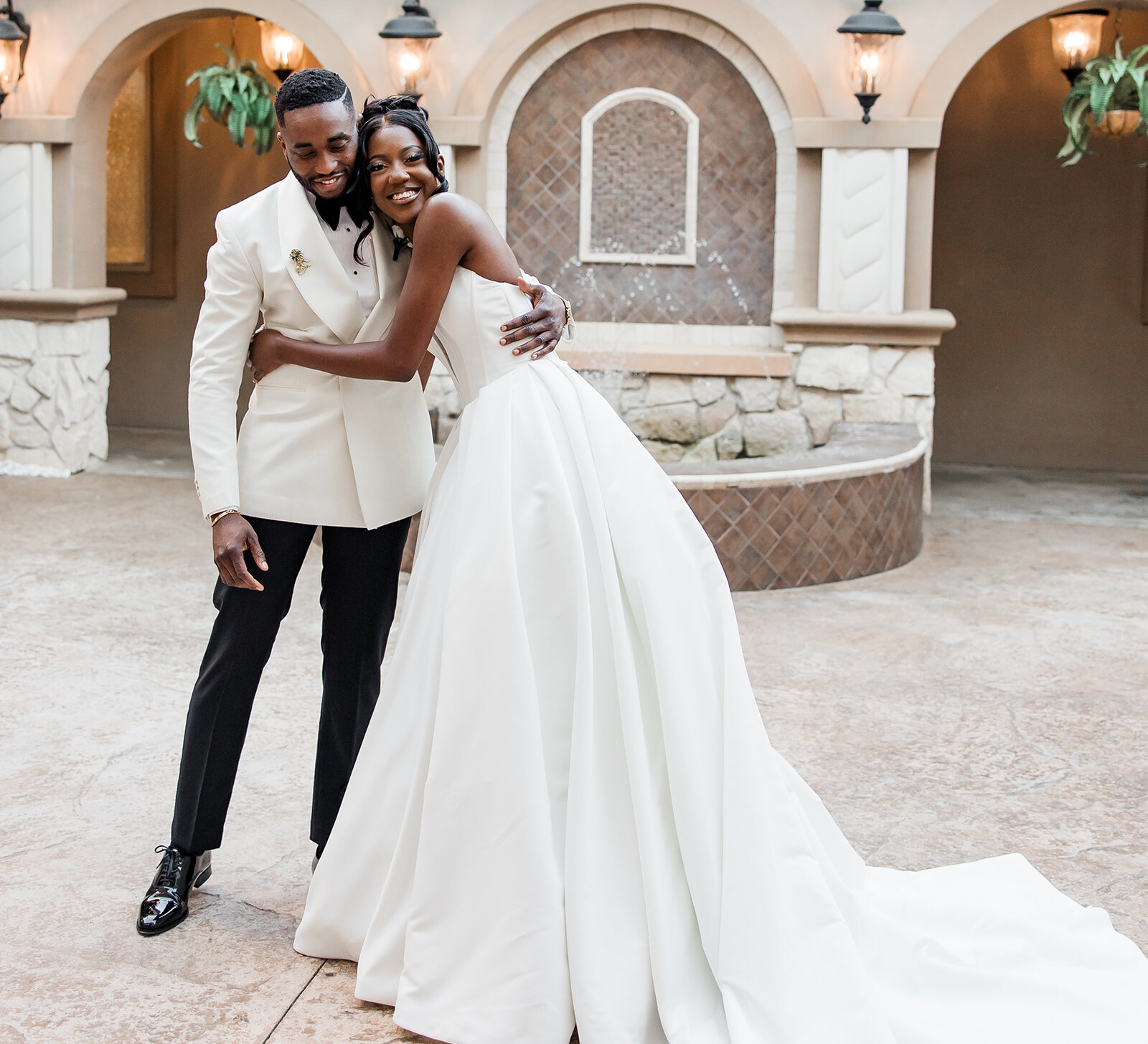 Bride and groom hugging and smiling in an elegant courtyard with a stone fountain, the bride in a flowing white gown and the groom in a cream jacket and black trousers