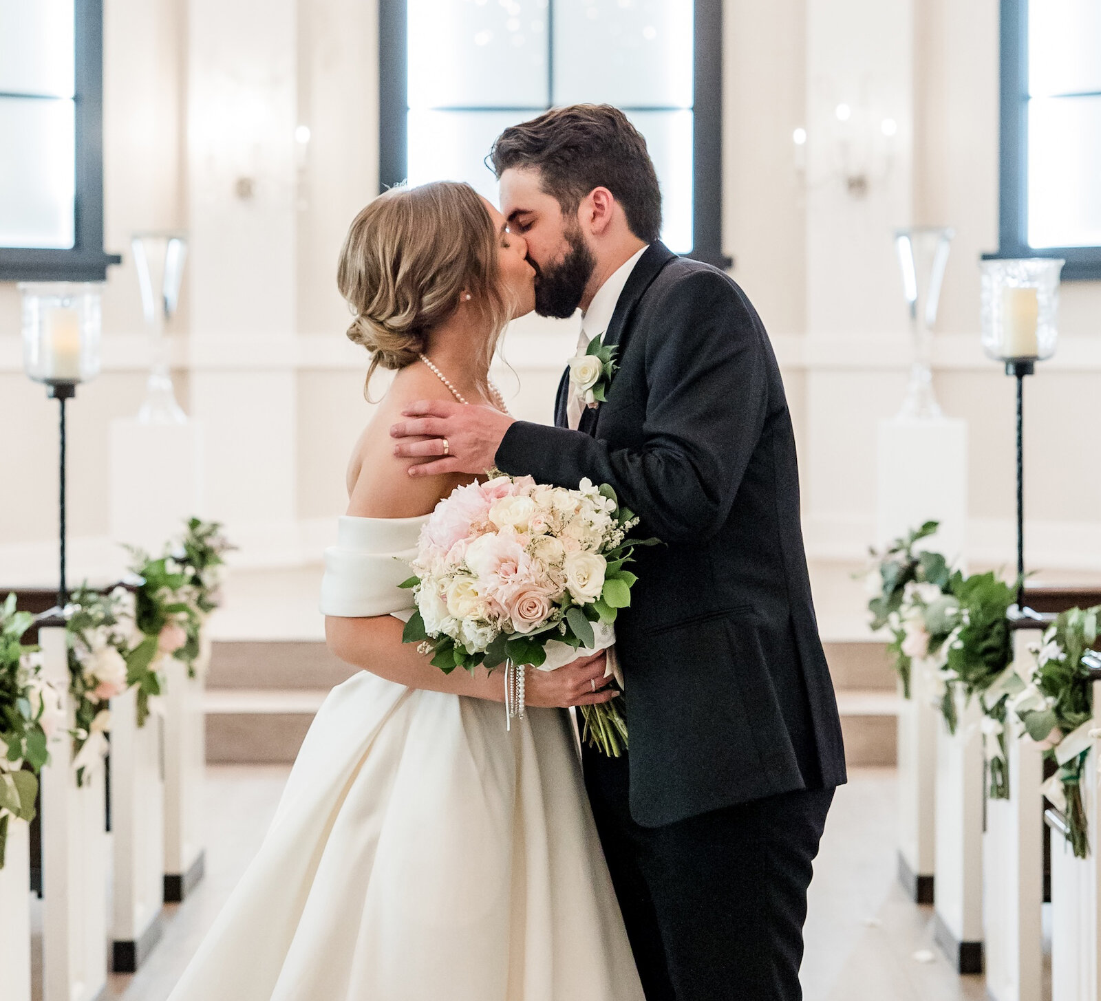 Romantic wedding kiss in a formal white chapel aisle under a crystal chandelier. The bride wears an off-the-shoulder satin ballgown and holds a large bouquet of pink and white roses, with the aisle decorated with greenery garlands.
