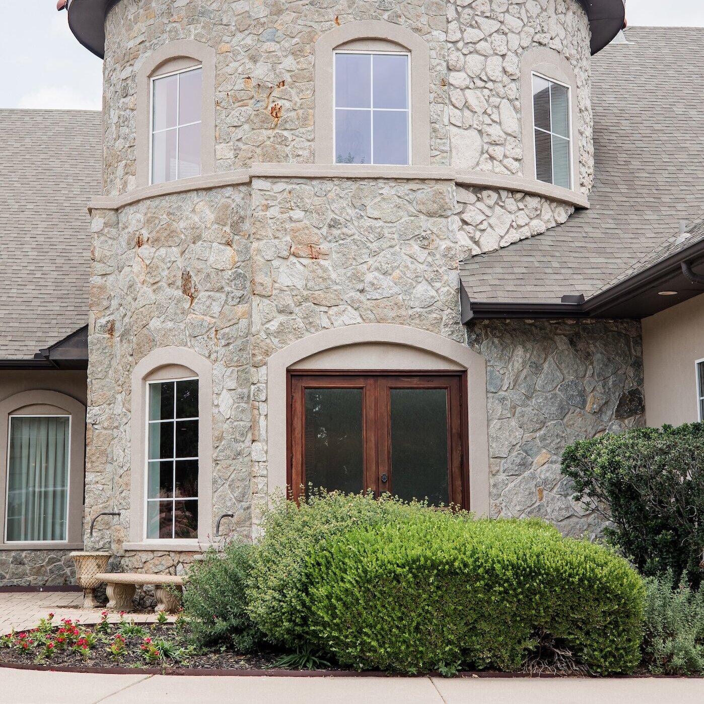 Exterior close-up of a round stone tower on a building, featuring a terracotta red tile roof and arched windows. The facade is constructed of rustic, light-colored quarry stone.