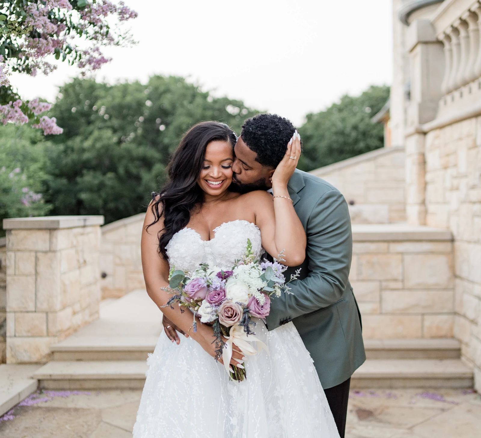 Bride and groom touching foreheads by a window, holding a white bouquet.