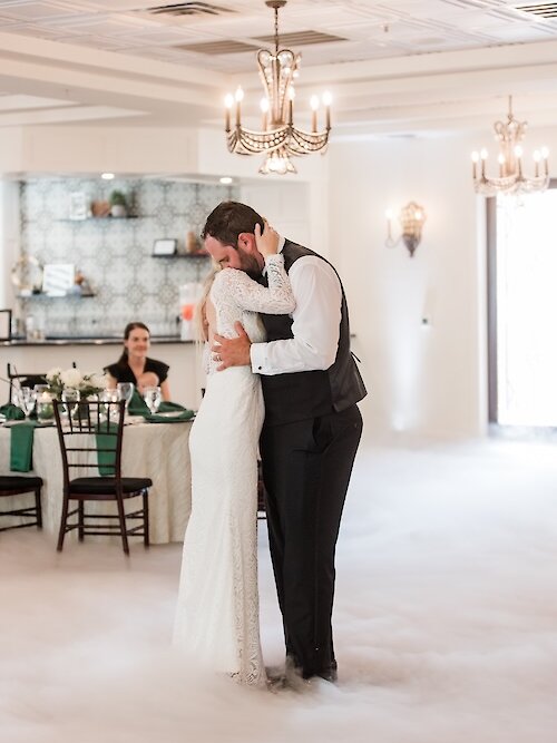 Romantic wedding first dance with the couple embracing tightly on a dance floor covered in low-lying fog (dancing on a cloud). The bride wears a long-sleeved lace gown, and the groom wears a vest and black trousers in a modern reception room.