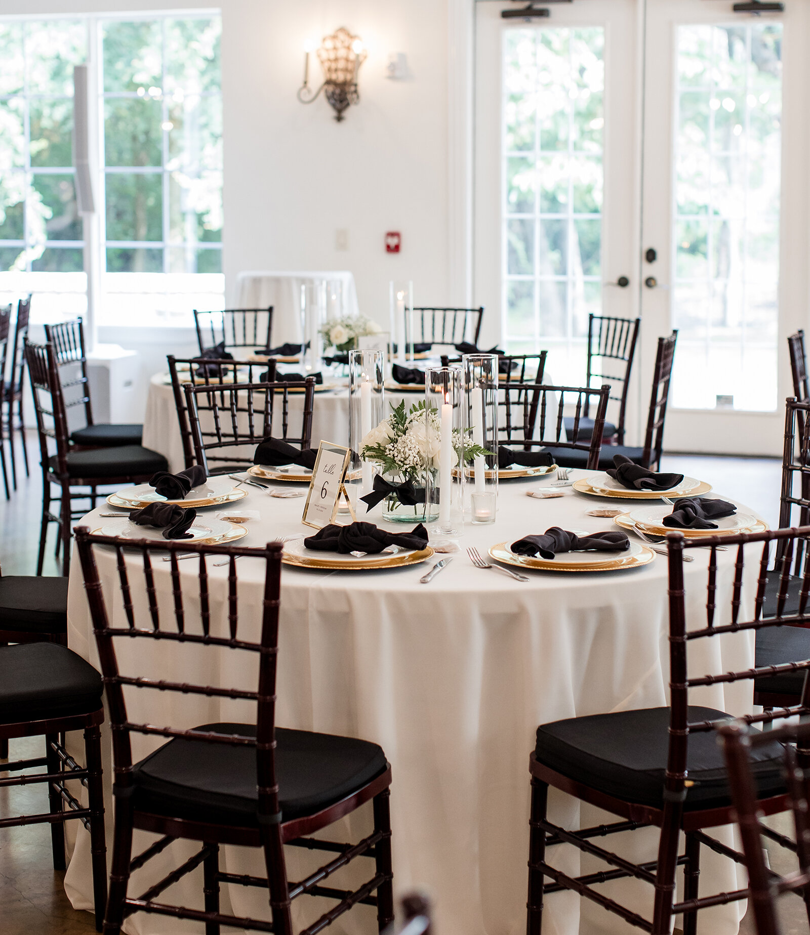 Elegant wedding reception setup featuring round tables with black and gold place settings, white floral centerpieces, and tall candles in a bright venue