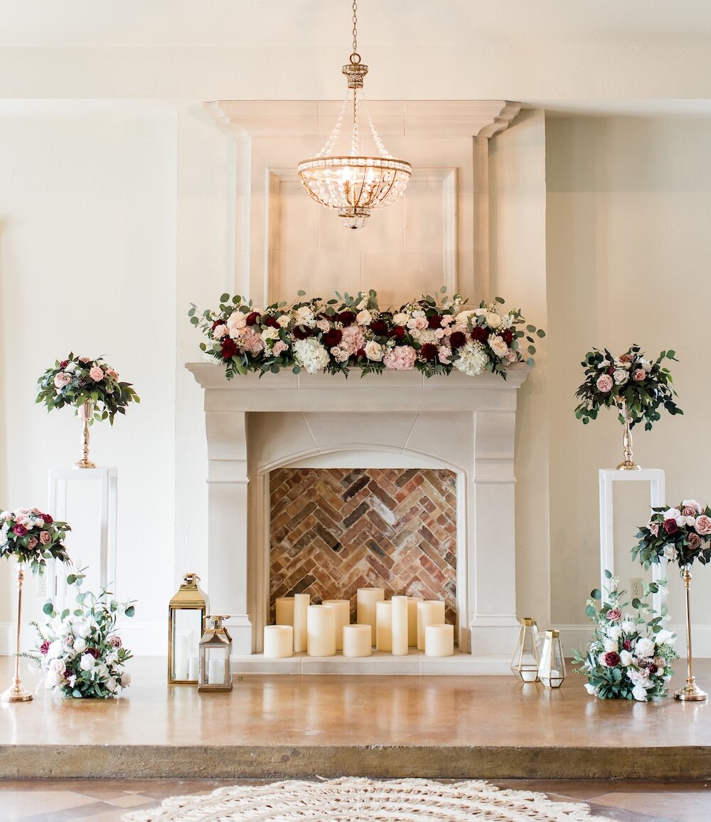 Elegant wedding ceremony altar setup featuring a white stone fireplace with a herringbone brick interior, decorated with a garland of deep red, burgundy, and white flowers, large pillar candles, and a gold beaded chandelier overhead.