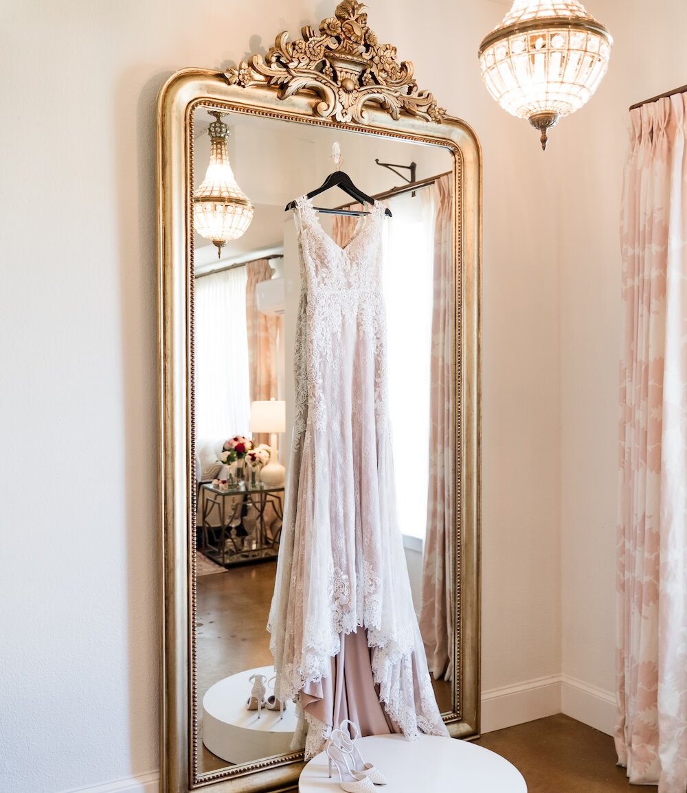 Luxury bridal details photo featuring a lace wedding dress with a layered blush pink lining hanging on a large, ornate gold-framed mirror in a brightly lit suite. White wedding shoes rest on a pedestal below.