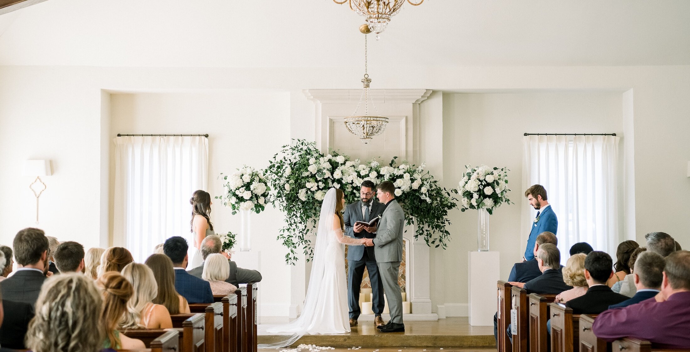 Bright and elegant indoor wedding ceremony inside a chapel with white walls and dark wooden pews. The couple is exchanging vows in front of a white floral garland and backdrop, beneath a classic gold chandelier.