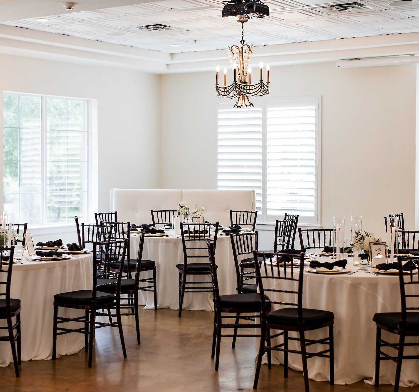Bright wedding reception room with round tables featuring white linens and contrasting black chiavari chairs. The tables are set with black napkins, gold charger plates, and simple white floral centerpieces.