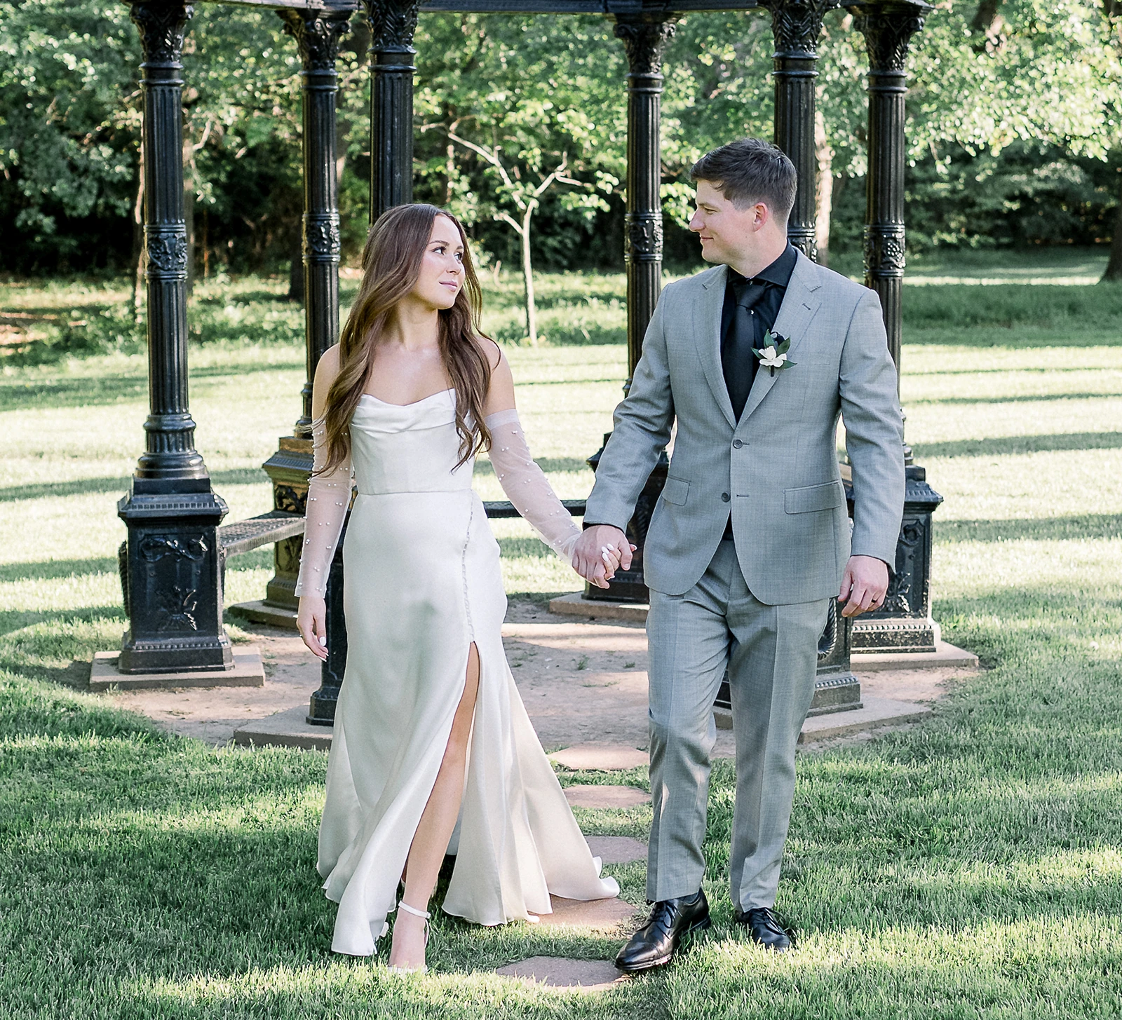 Bride and groom walking hand in hand in front of a decorative gazebo in a garden, looking at each other during their wedding portraits.