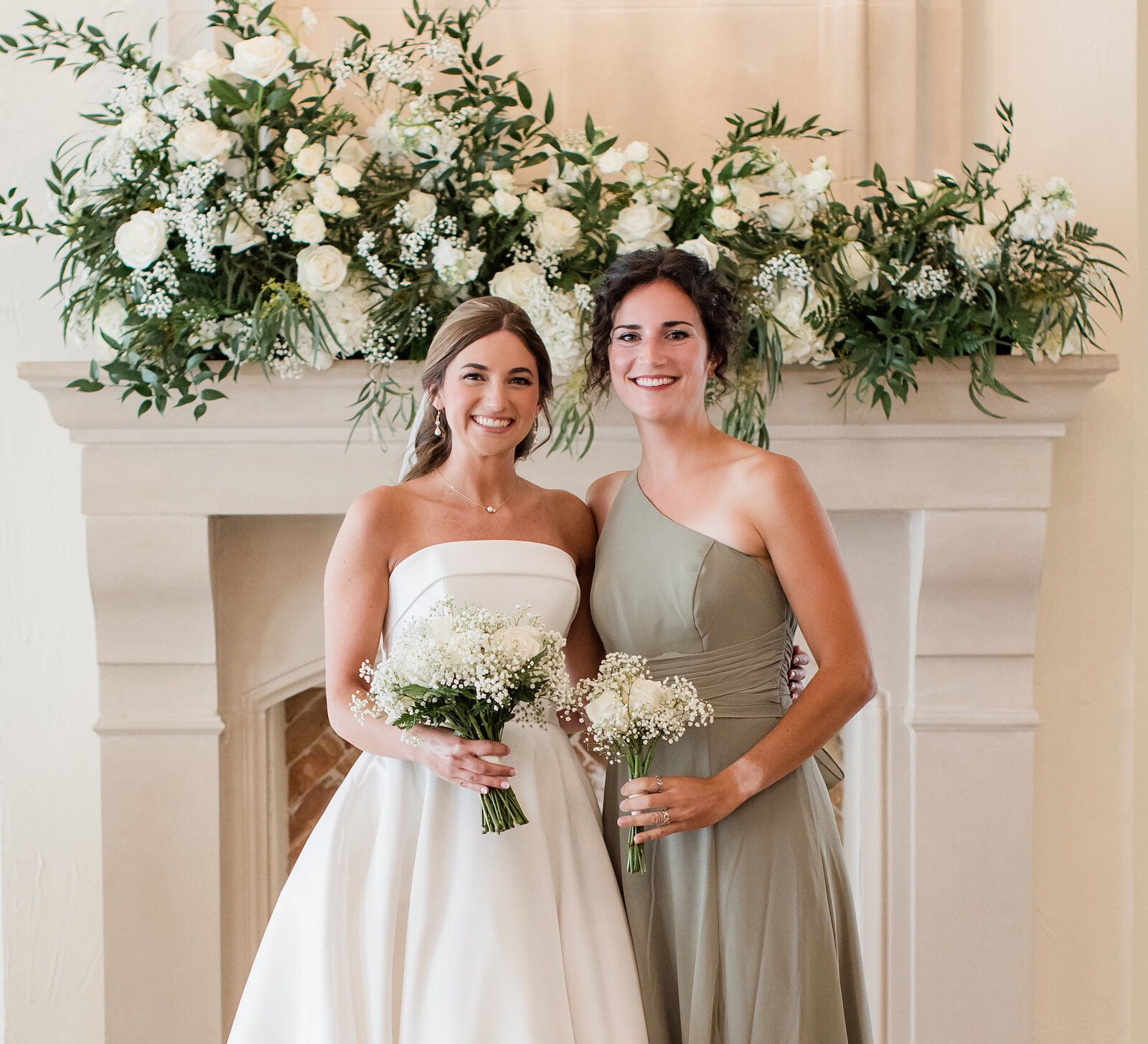 Bride and bridesmaid posing in front of a white stone fireplace decorated with a sweeping white and green floral garland. The bride wears a strapless satin ballgown and the bridesmaid wears an olive green one-shoulder dress.
