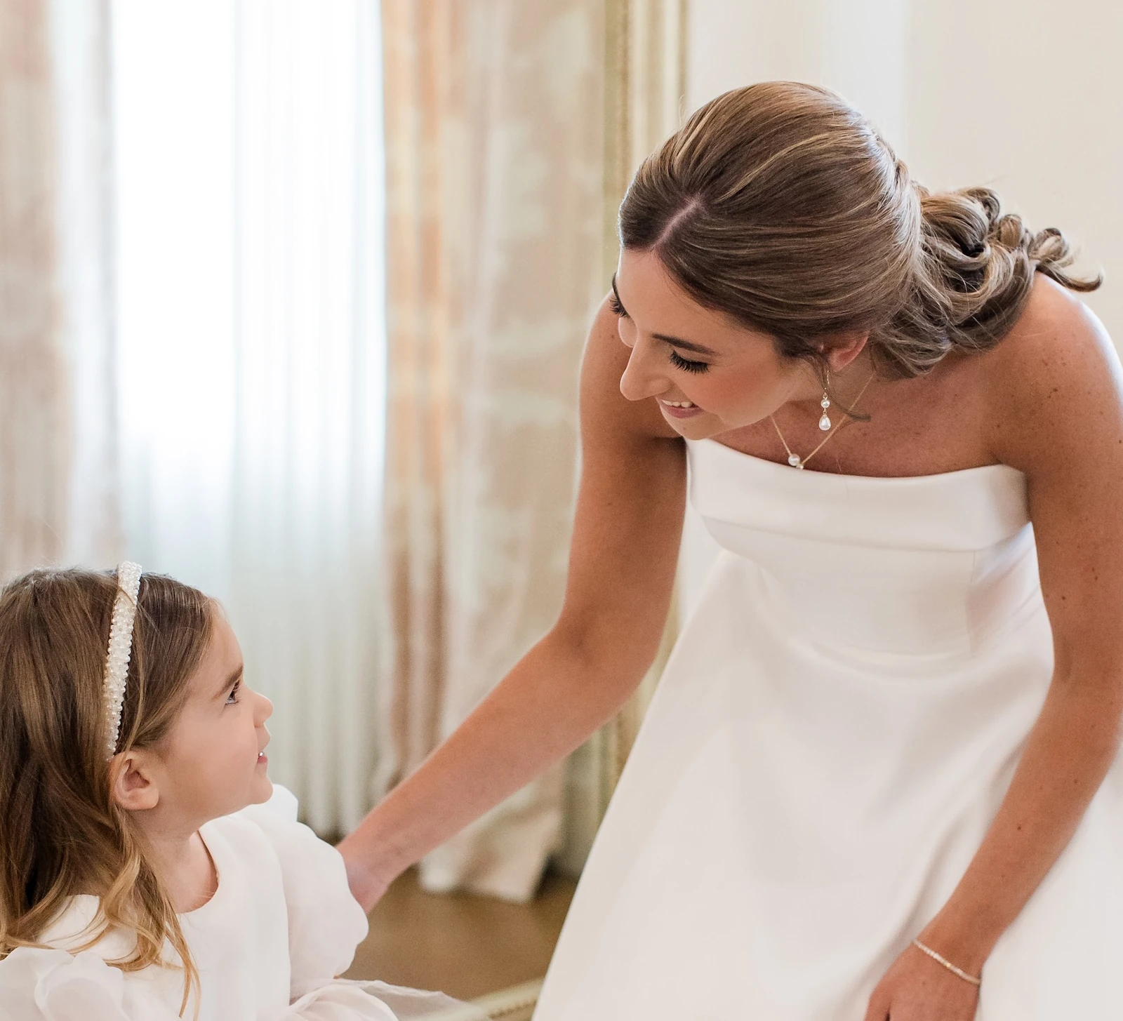 Sweet moment of the bride smiling down at the flower girl in a bright bridal suite. The flower girl wears a puffy-sleeved white tulle dress and the bride wears a strapless white satin ballgown.