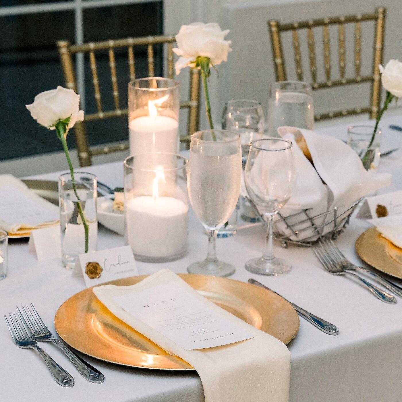 An elegant wedding dinner table setting featuring a gold charger plate, menu card, large pillar candles, and single white roses.
