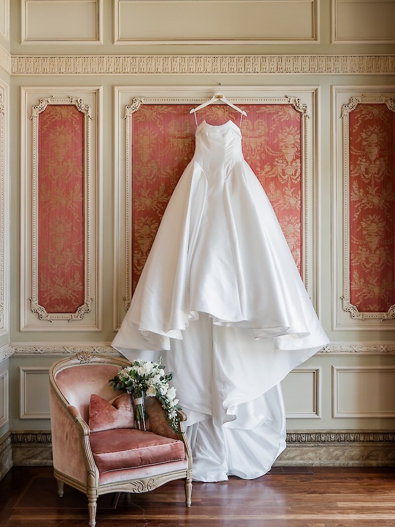 Bridal gown hanging on ornate wall panel with white flower bouquet in adjacent chair