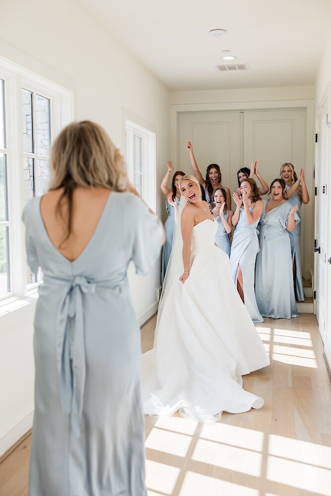 Excited bride revealing dress to mother with cheering bridal party behind her