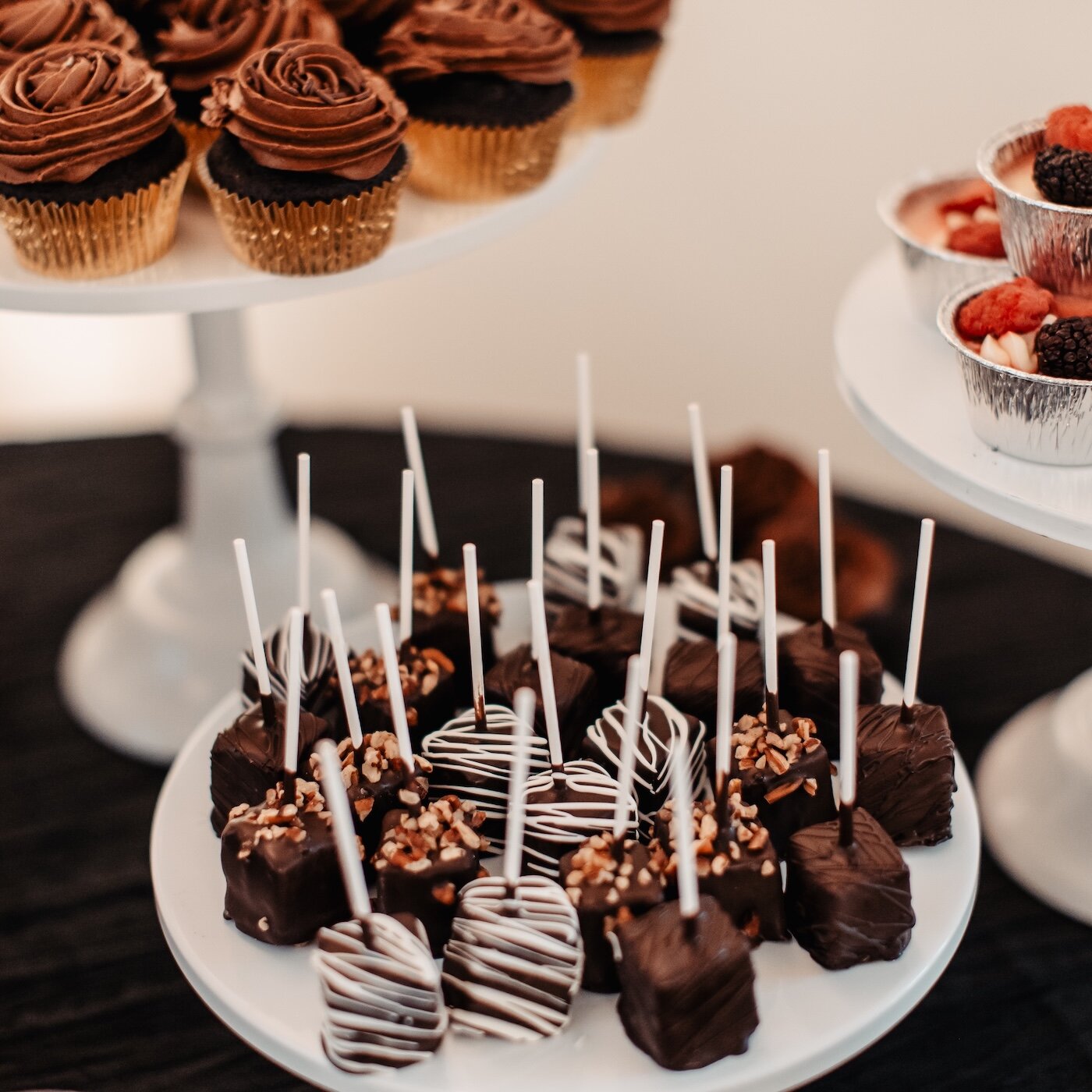 A wedding dessert table display featuring chocolate cake pops with white sticks and chocolate cupcakes on white pedestal stands.