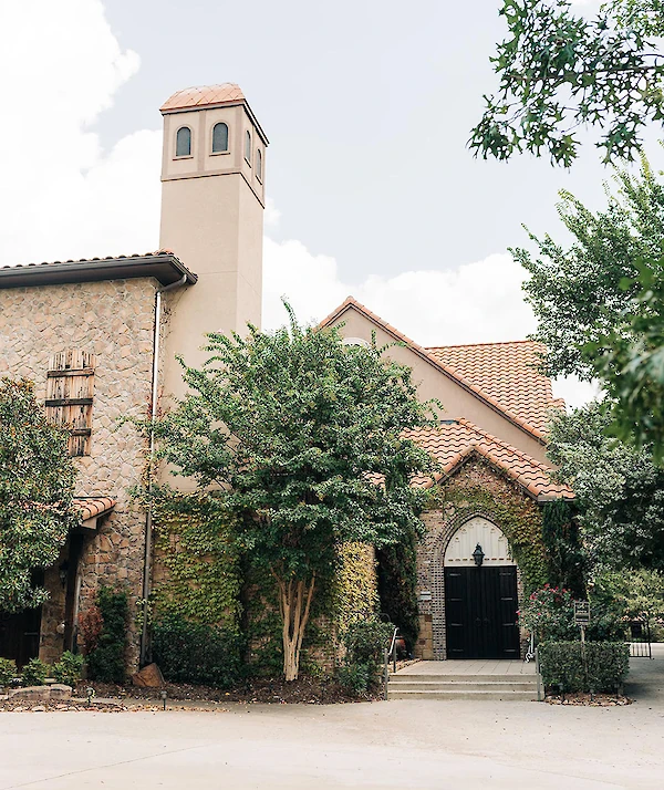 A charming stone building with terracotta roof tiles, arched windows, a small tower, and string lights hanging above a landscaped staircase and garden.