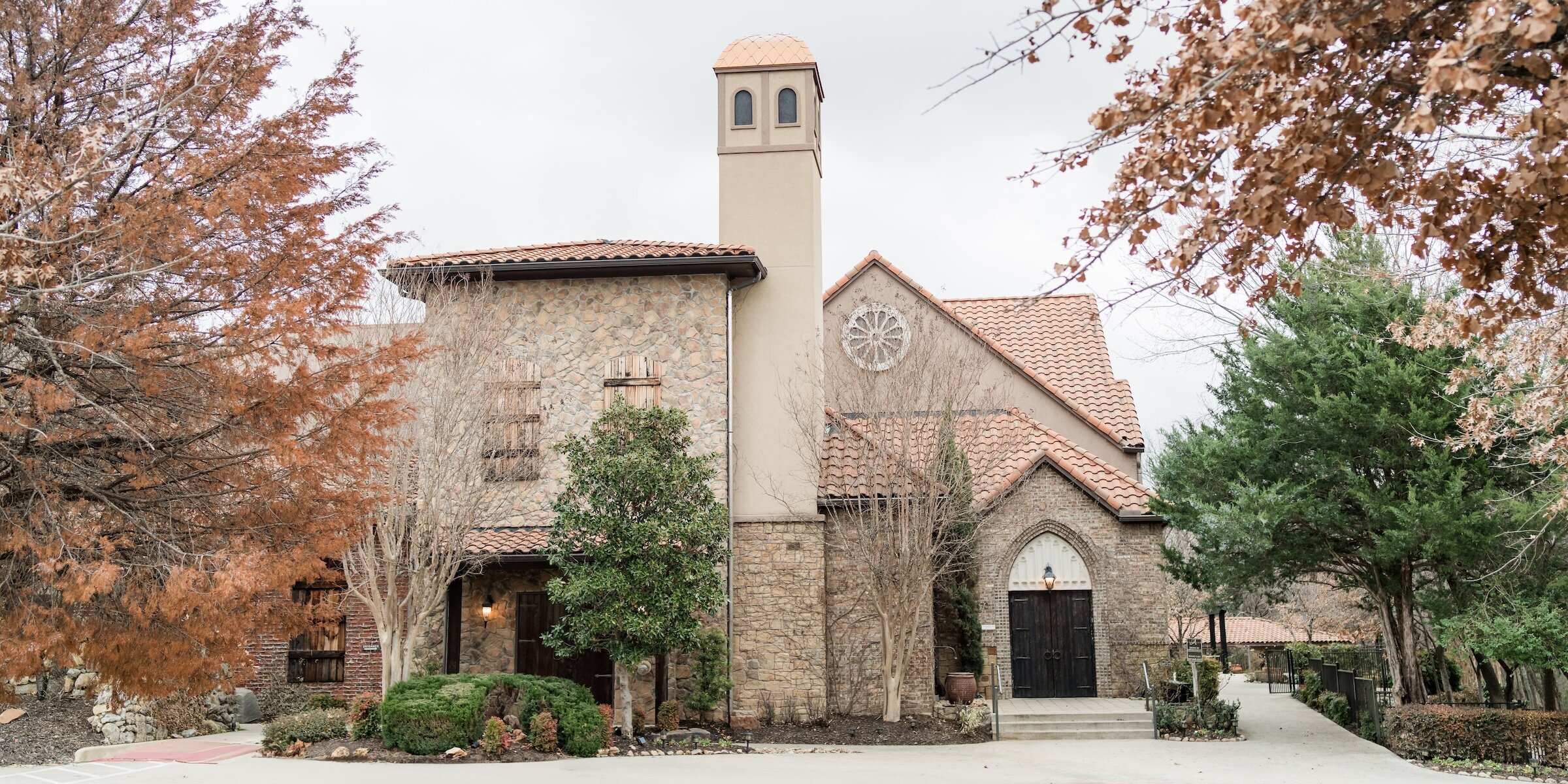 Exterior of a rustic Mediterranean-style wedding chapel featuring a stone facade, terracotta tile roof, and a tall, square bell tower. The scene includes autumn or winter trees with copper-colored leaves.