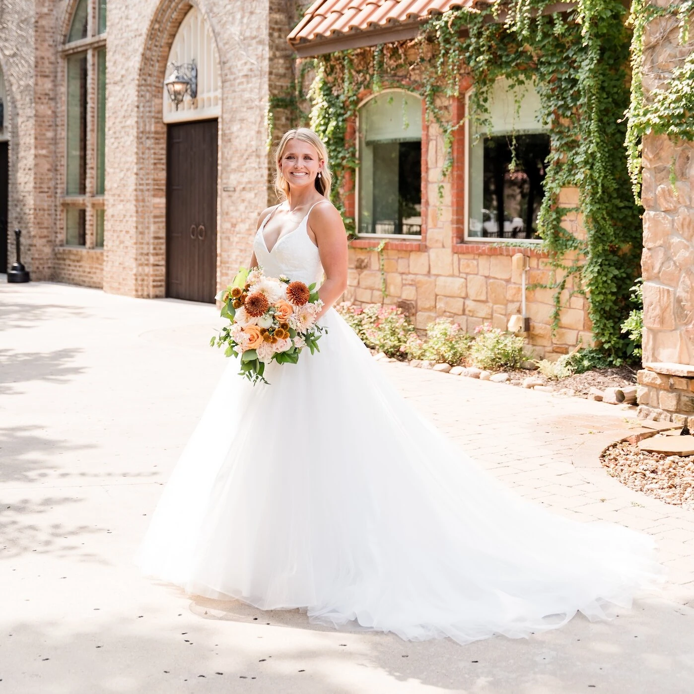 Bridal portrait of the bride smiling in front of a rustic stone and brick chapel with a terracotta tile roof, featuring abundant ivy. She wears a white ballgown with a plunging neckline and holds a fall-inspired bouquet with orange, peach, and white flowers.