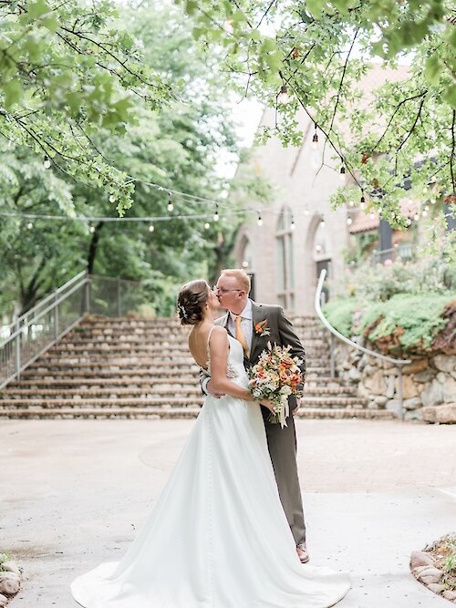 Romantic wedding portrait of the bride and groom kissing outdoors under a canopy of green trees and string lights. The background features a large stone staircase leading up to a classic stone venue. The bride wears a sleek, backless satin A-line gown and holds an orange and white bouquet.