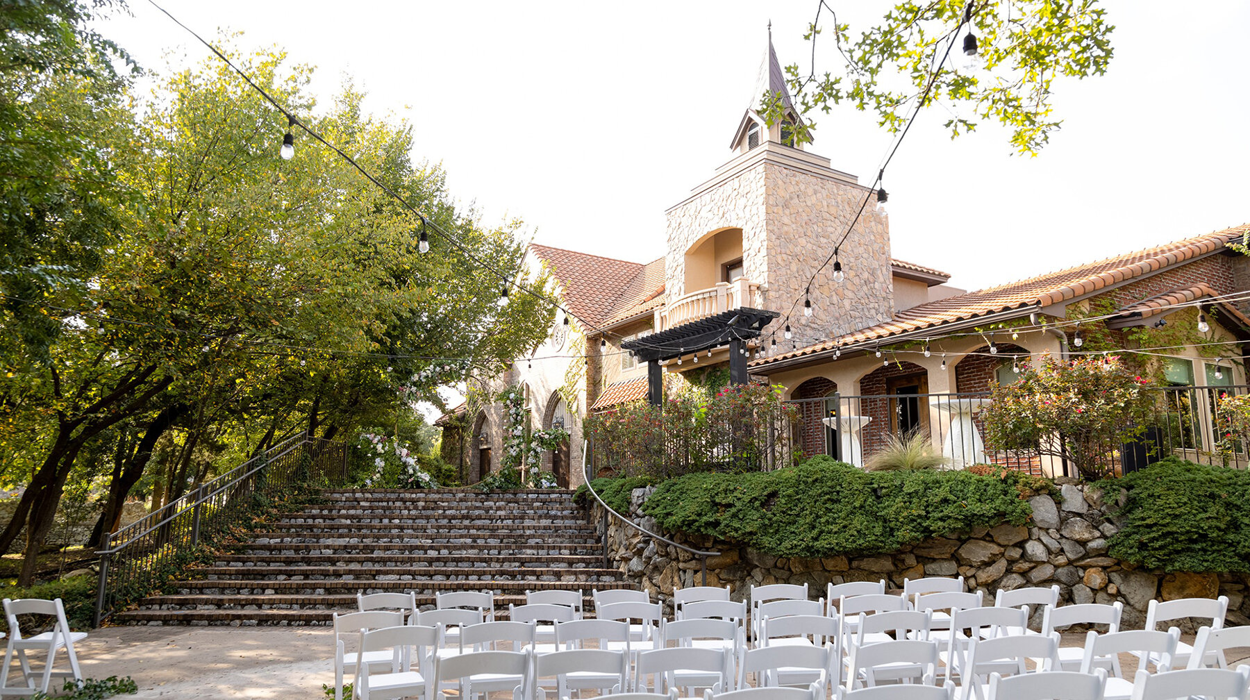 Outdoor wedding ceremony setup with white chairs facing a large stone staircase at a Mediterranean-style venue decorated with string lights and lush greenery.