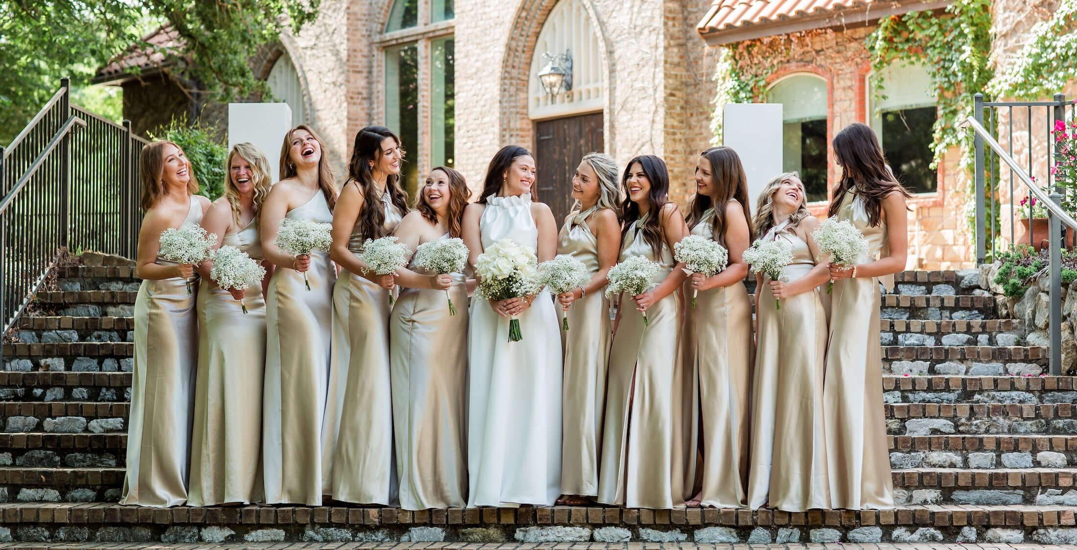 Joyful bridal party portrait with the bride and eleven bridesmaids laughing on a wide, tiered stone staircase. The bridesmaids wear matching champagne satin slip dresses and carry bouquets of white baby's breath, with a gothic-style stone chapel in the background.