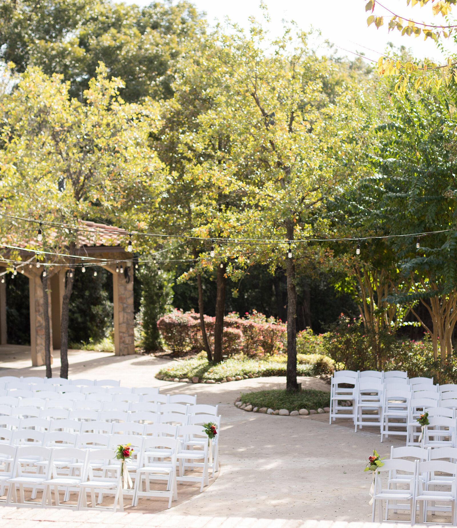 Outdoor garden wedding ceremony setup with rows of white chairs, string lights overhead, and trees with early fall foliage