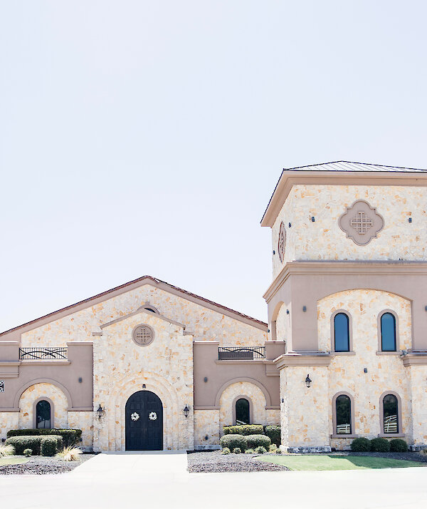 Front view of a light stone building with arched windows, a tall square tower, dark double doors, and landscaped greenery under a bright, cloudy sky.