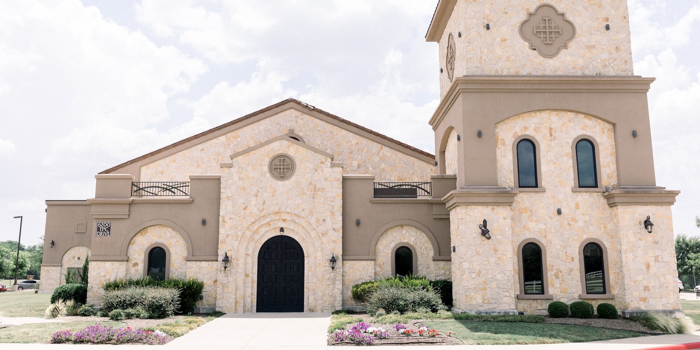 Exterior of a large Mediterranean-style building with a prominent square tower, featuring a facade of light beige stucco and rough-cut stone. The entrance has large, decorative black metal doors and arched windows.