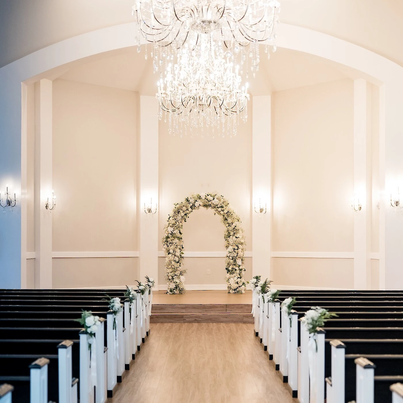 Elegant white wedding chapel interior with an arched altar featuring a tall white floral and greenery arch. The aisle has a light wood floor, leading to dark wood pews, all illuminated by a magnificent cascading crystal chandelier.