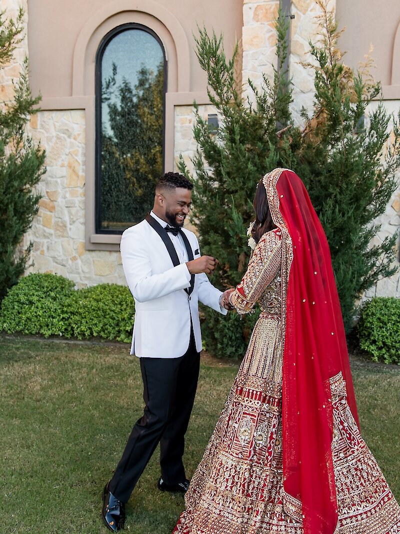 Vibrant cultural wedding portrait of a couple holding hands on a green lawn outside a stone venue. The groom wears a white tuxedo jacket, and the bride wears a traditional, heavily embroidered red and gold bridal lehenga with a matching red dupatta veil.