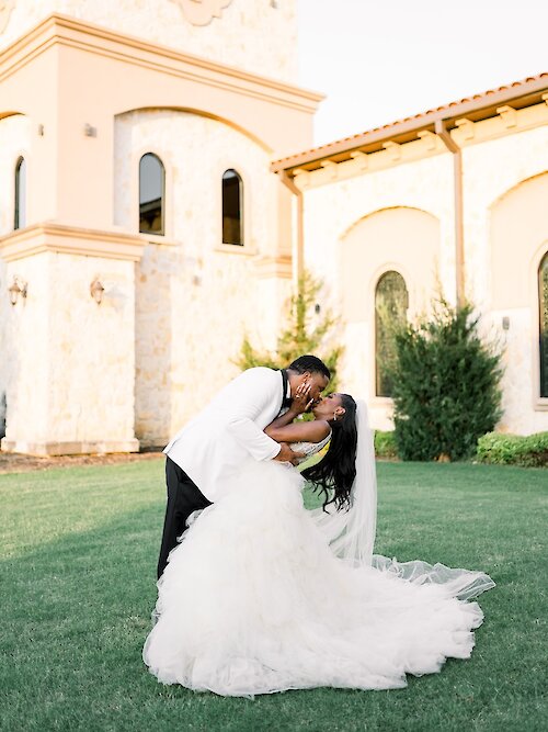 Dramatic wedding portrait of a Black couple kissing and dipping on a sunny green lawn in front of a Mediterranean-style stone chapel. The groom wears a white tuxedo jacket, and the bride wears a voluminous, tiered white tulle ballgown.