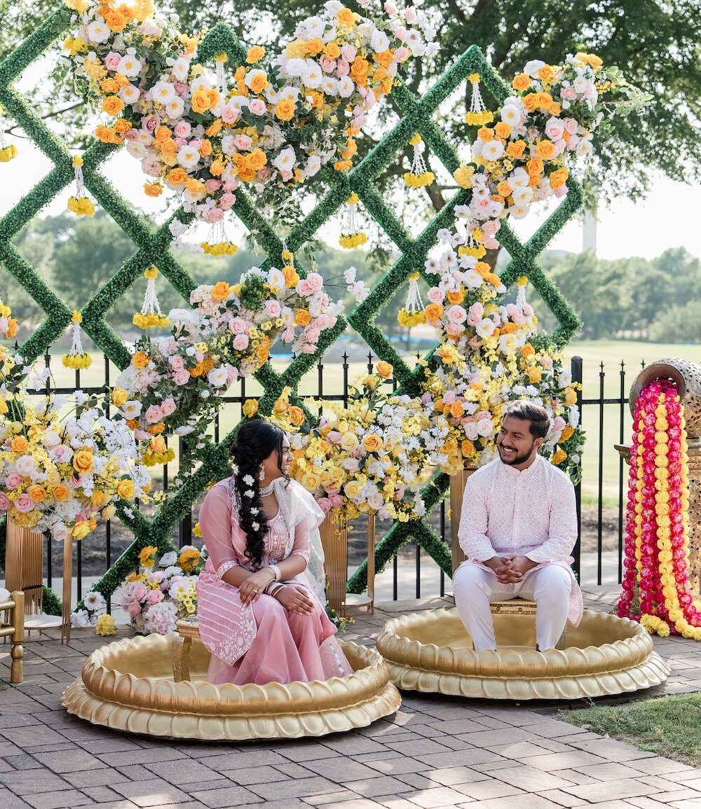 The bride and groom sit in golden lotus bowls for an Indian wedding ceremony in front of a green lattice backdrop decorated with yellow and pink flowers.
