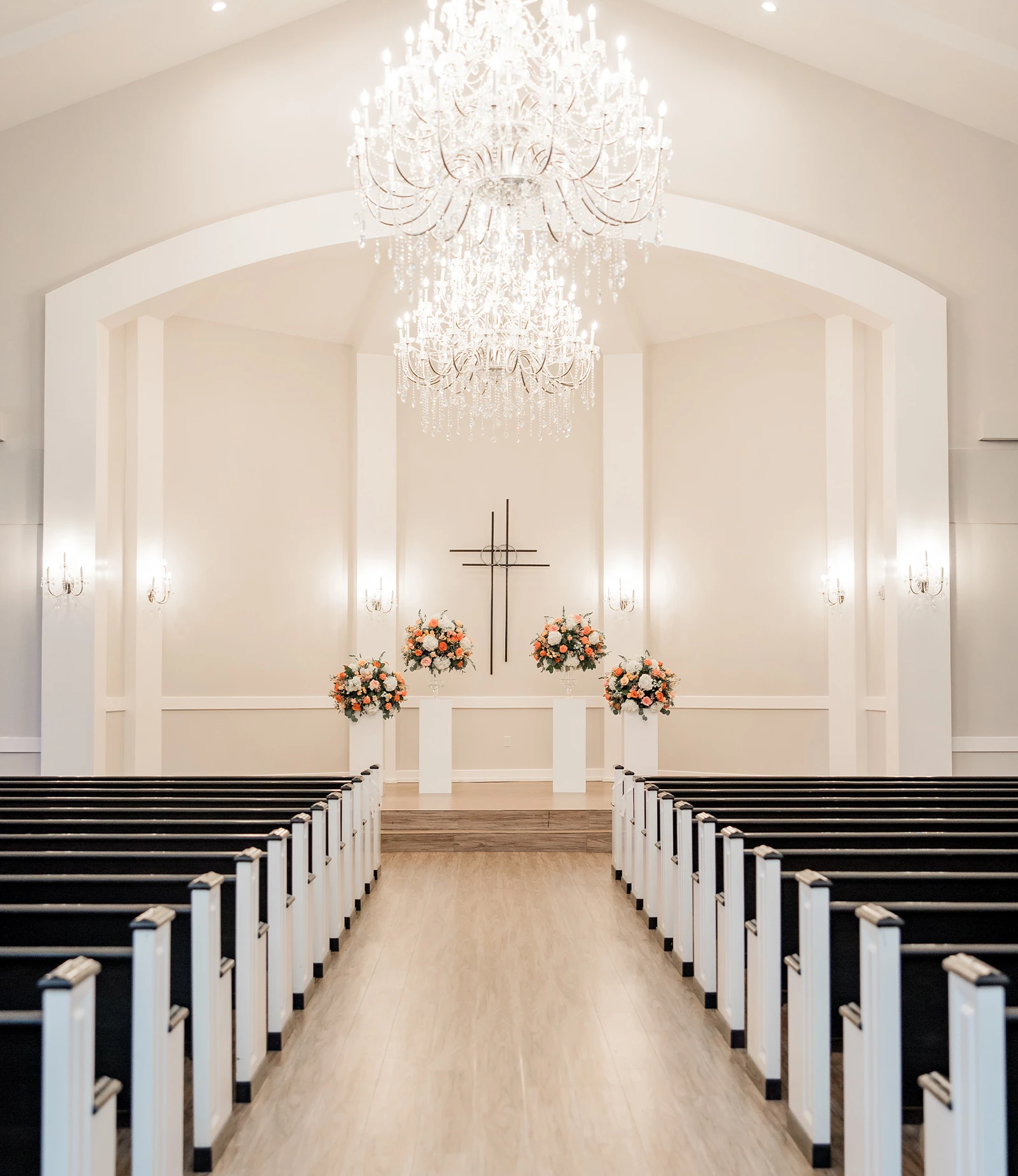 The elegant interior of a wedding chapel featuring a long aisle, dark pews, white floral arrangements, and a tiered crystal chandelier.