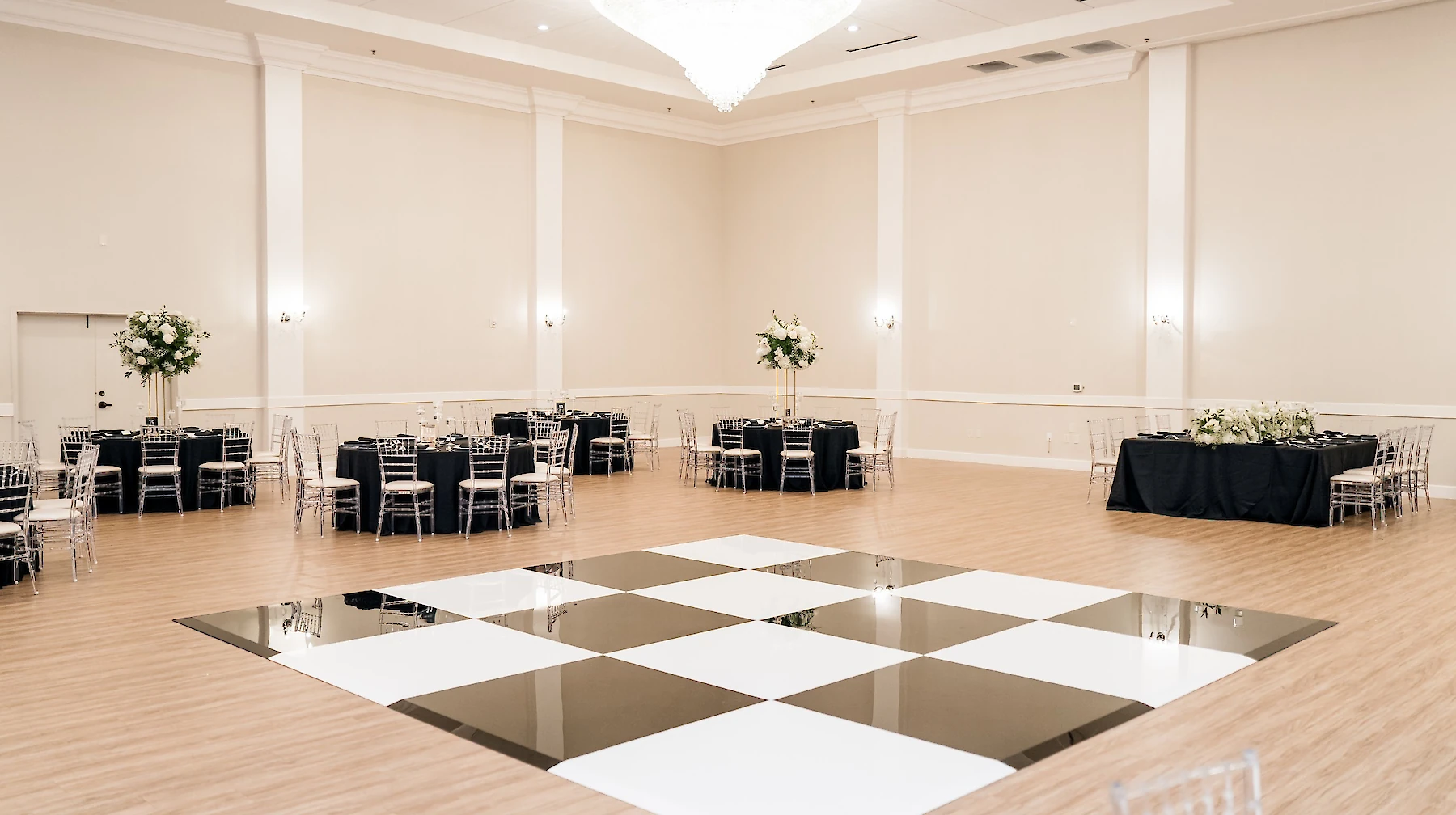 Elegant wedding ceremony inside a bright, modern chapel with a vaulted ceiling and crystal chandelier. The couple exchanges vows at the altar, which is framed by two large white floral arches and a cross.