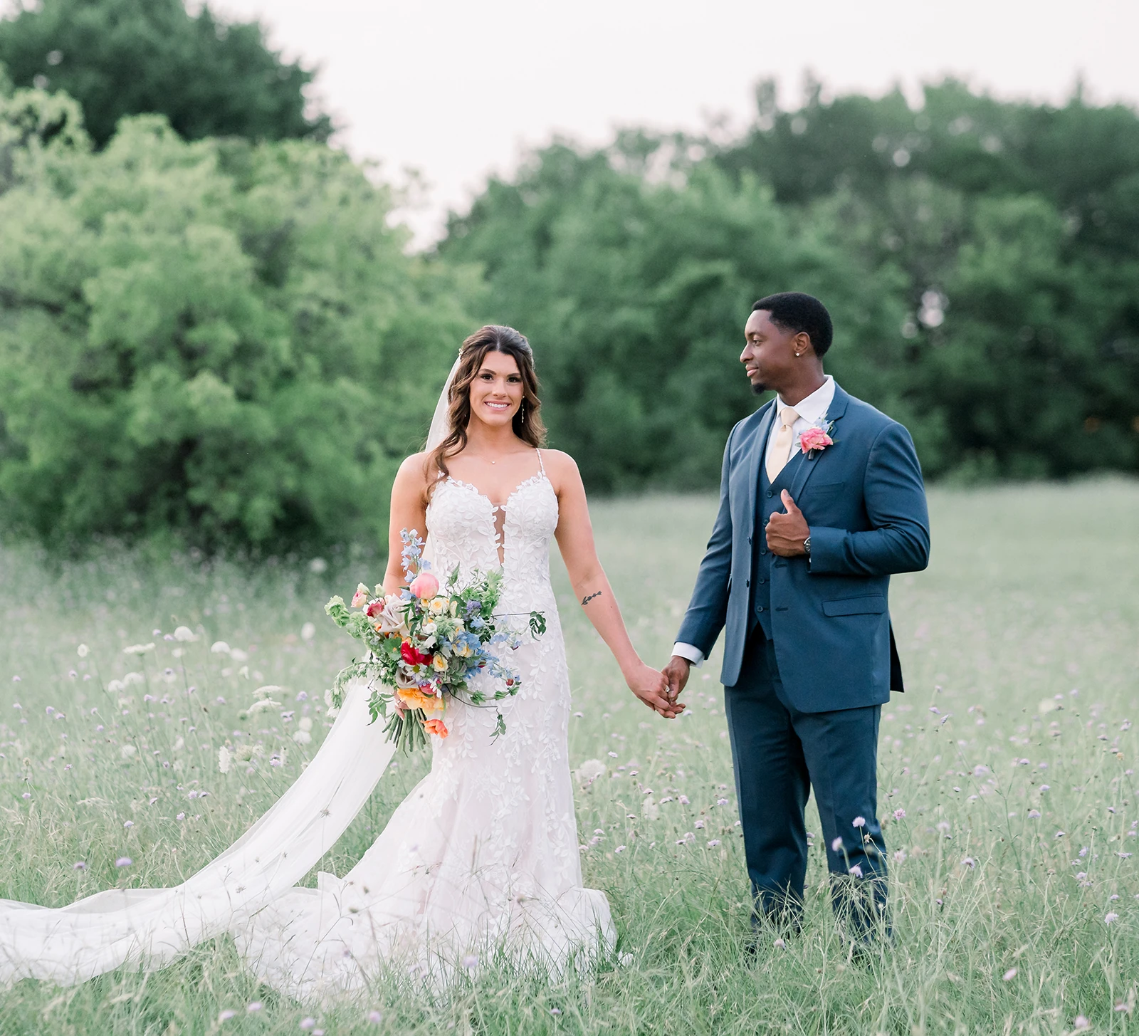 Bride and groom holding hands in a wildflower field, with the bride wearing a lace gown and colorful bouquet and the groom in a blue suit