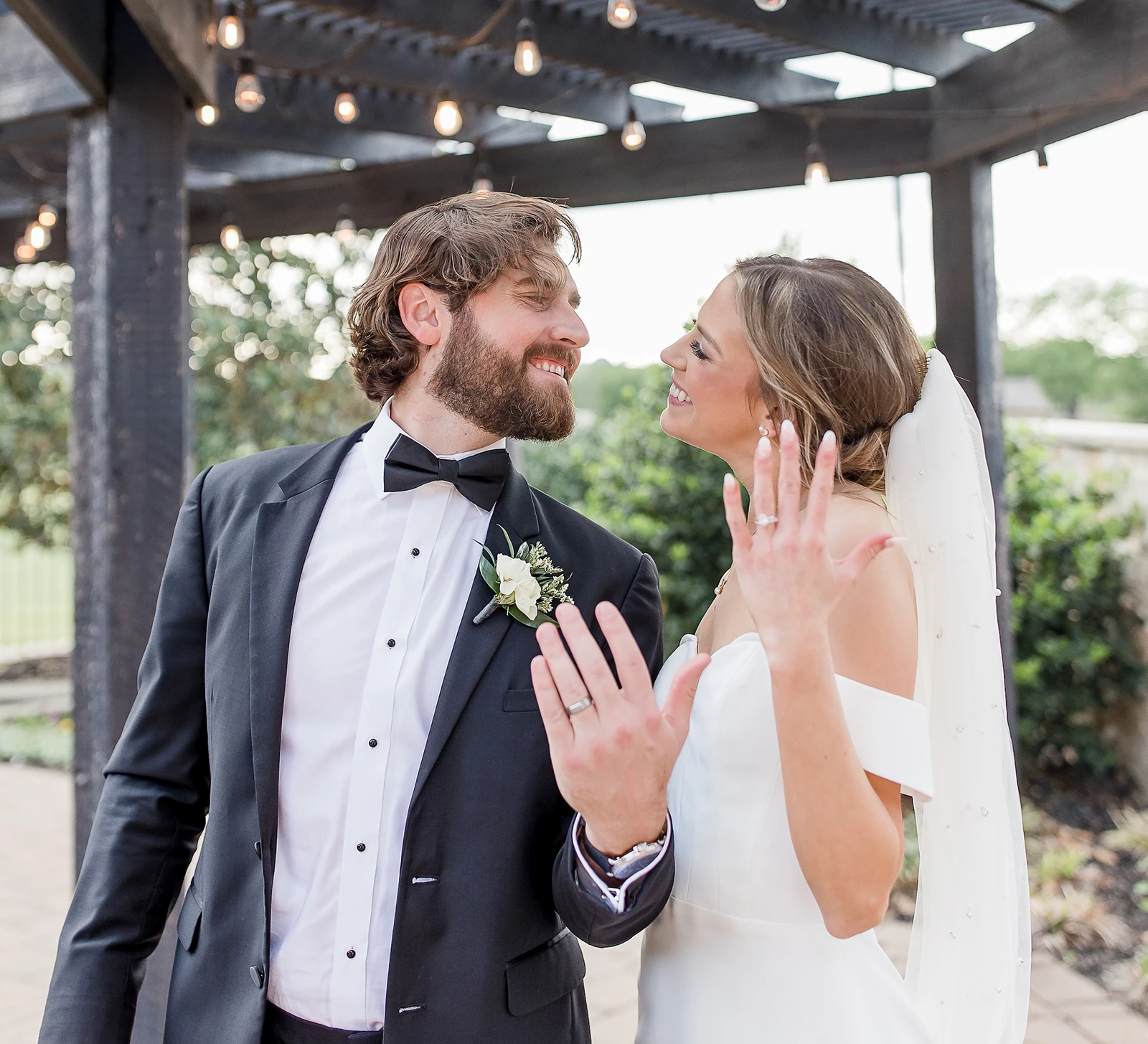 Joyful newlywed portrait showing off their wedding rings under a dark wooden pergola decorated with string lights. The bride wears a sleek, off-the-shoulder gown, and the groom wears a black tuxedo.