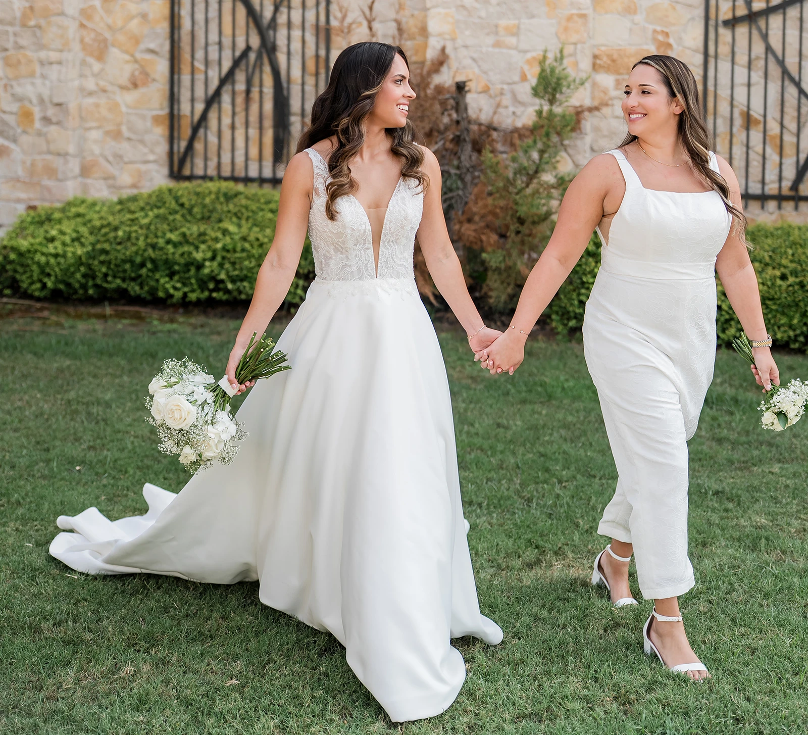 Two brides holding hands and walking together outdoors on their wedding day, wearing white outfits and carrying white bouquets