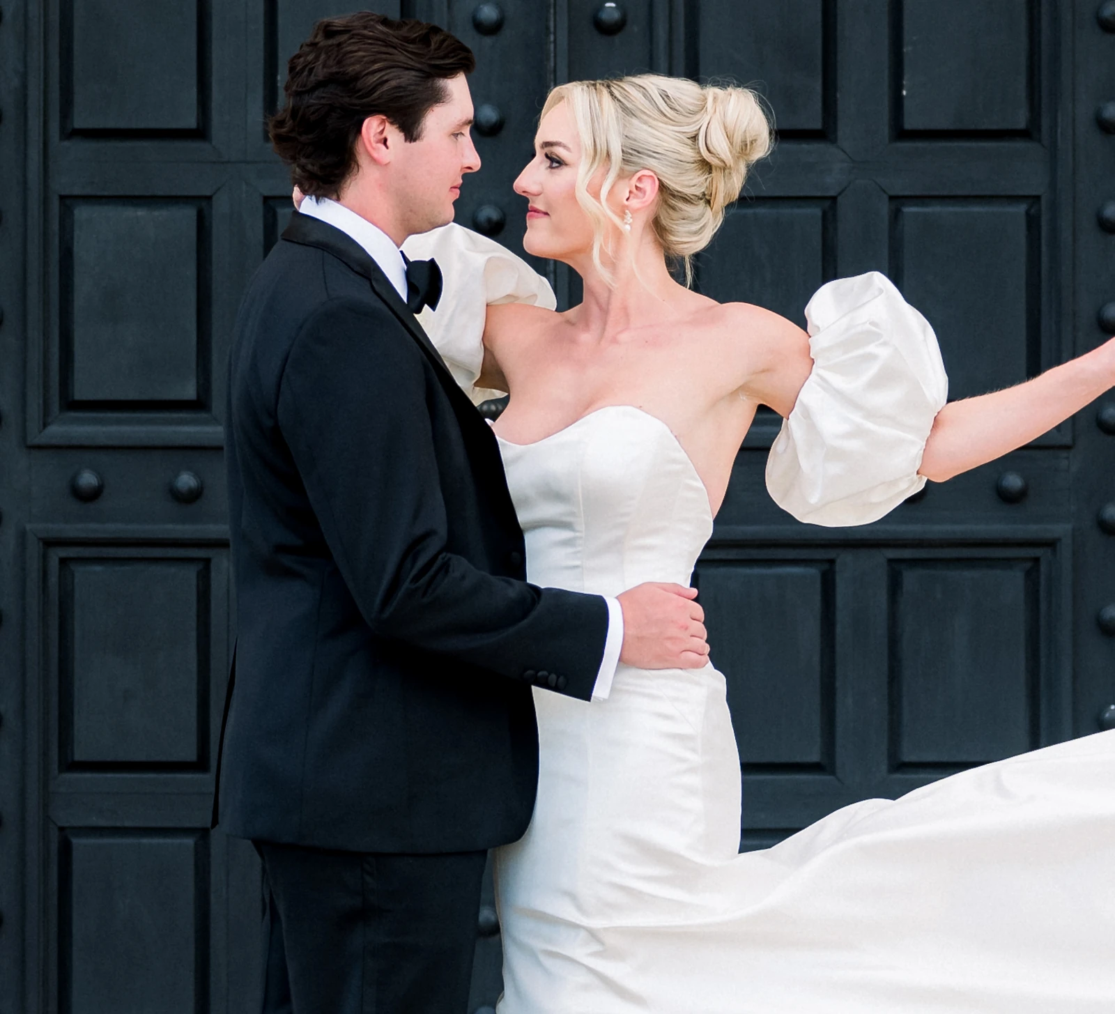 Bride and groom embracing and looking at each other in front of large black doors, with the bride wearing a strapless gown with puff sleeves.
