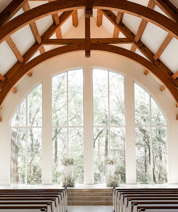 “Front view of a light stone chapel with tall arched wooden doors, narrow arched windows, and a circular window above, surrounded by landscaped greenery and trees.”