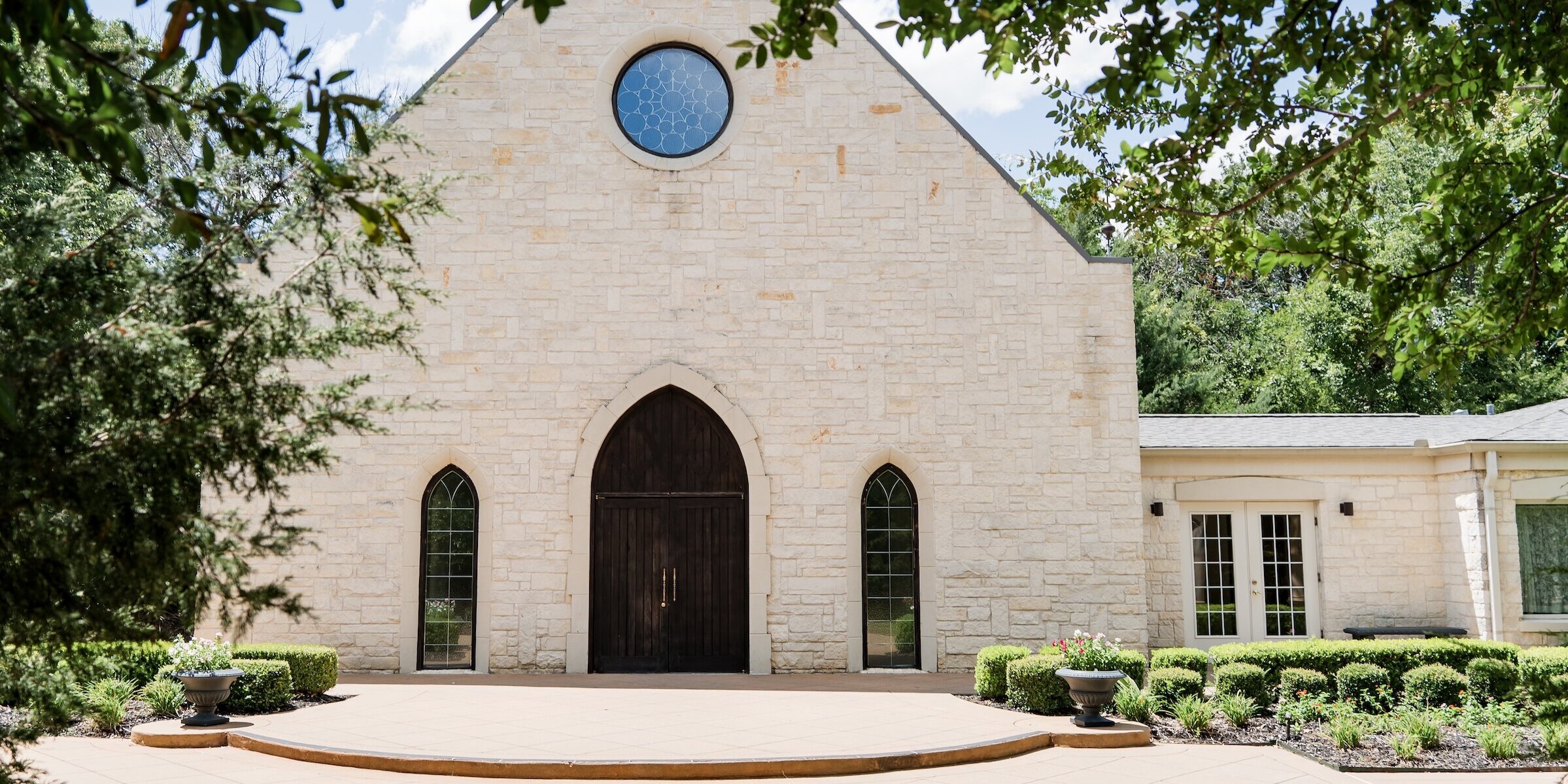 Exterior of a quaint white stone wedding chapel with an A-frame roof, dark wood arched doors and windows, and a central circular stained-glass window, framed by green trees on a sunny day.