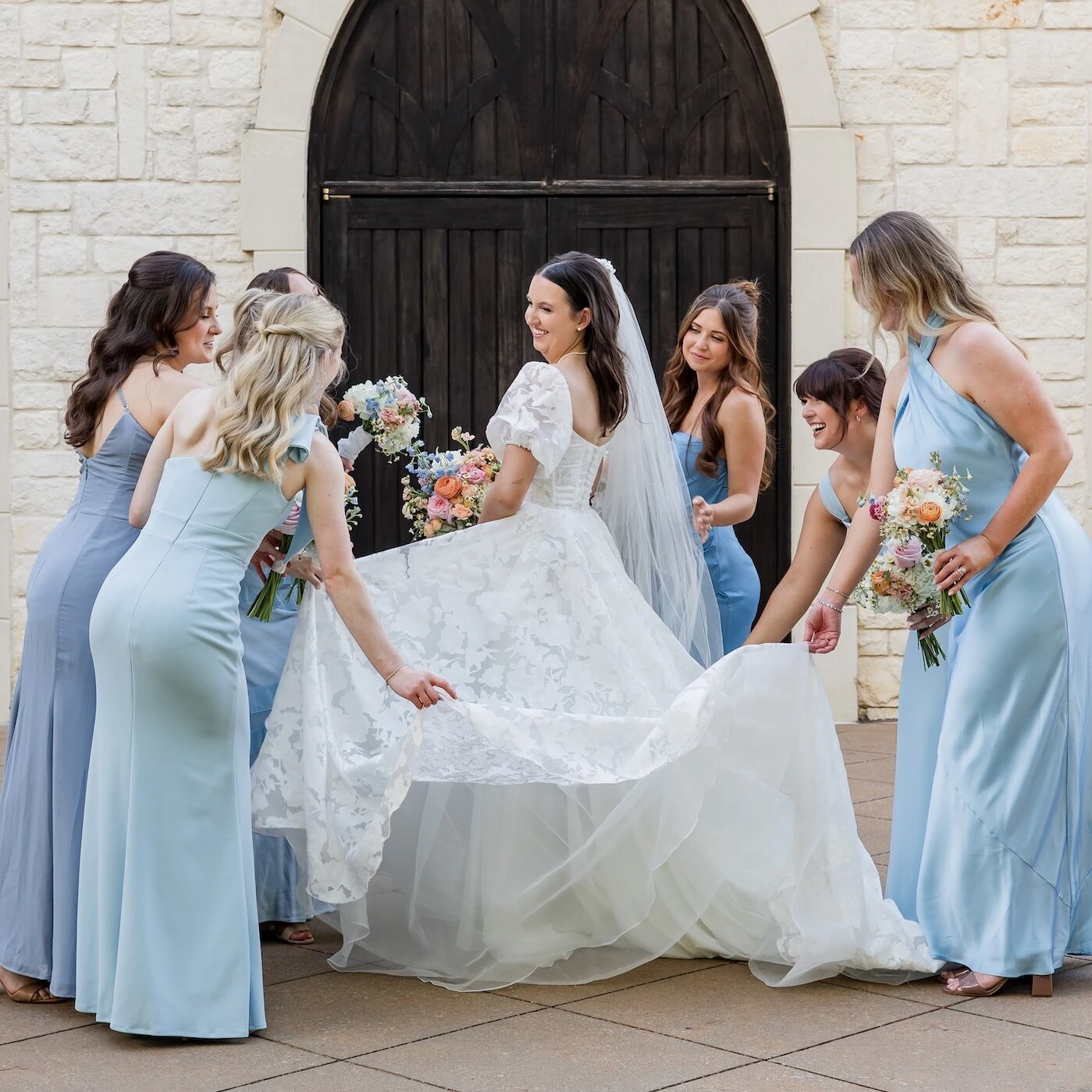 Playful bridal party portrait with the bride and her five bridesmaids adjusting and admiring the long train of her lace and tulle gown outside a white stone chapel with dark arched doors. The bridesmaids wear coordinating light blue satin dresses.
