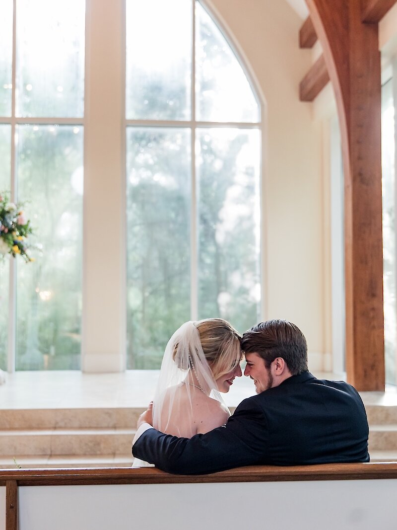 Intimate profile portrait of the bride and groom touching foreheads while seated in a wooden pew inside a sunlit chapel. The background features a large arched window overlooking the outdoors and exposed wooden architectural beams.
