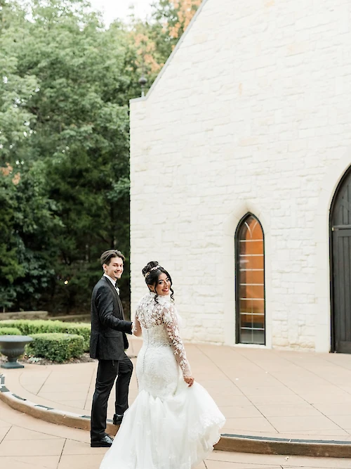 Joyful newlywed portrait of the bride and groom walking away from a white stone chapel, holding hands, with the bride looking back and smiling. The bride wears a long-sleeved lace wedding gown with a mermaid skirt.