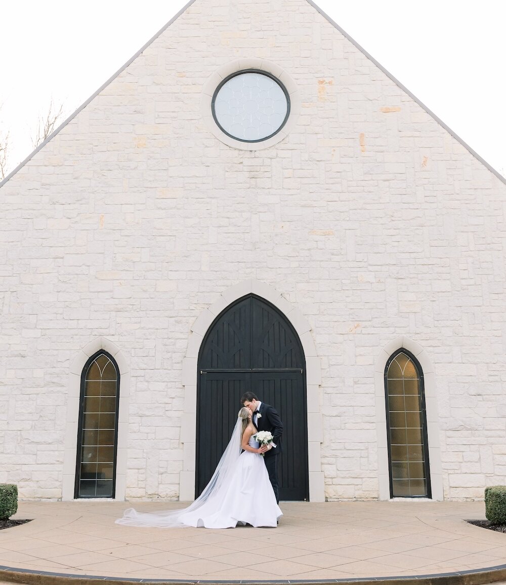 The bride and groom kiss in front of a white stone chapel's black arched doors, with the bride's long veil dramatically trailing behind her.