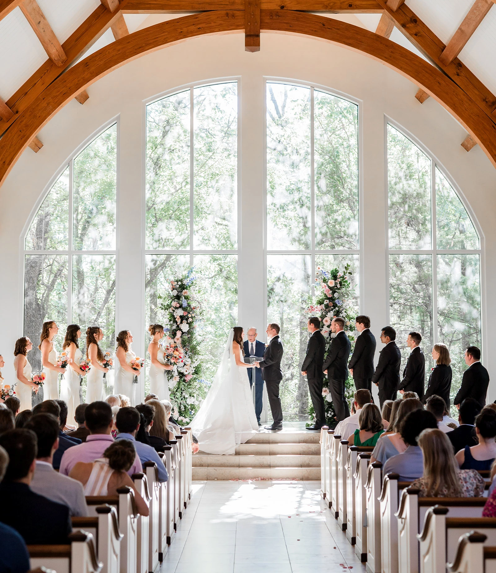 Joyful and energetic portrait of the groom and seven groomsmen in black tuxedos laughing and celebrating outside a white stone chapel with dark arched doors.