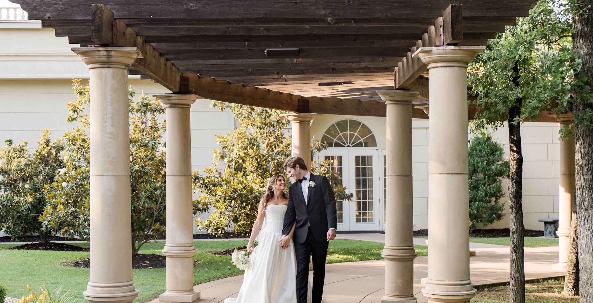 The bride and groom walk hand-in-hand down a paved walkway under a wooden pergola supported by thick stone columns outside a light-colored venue.
