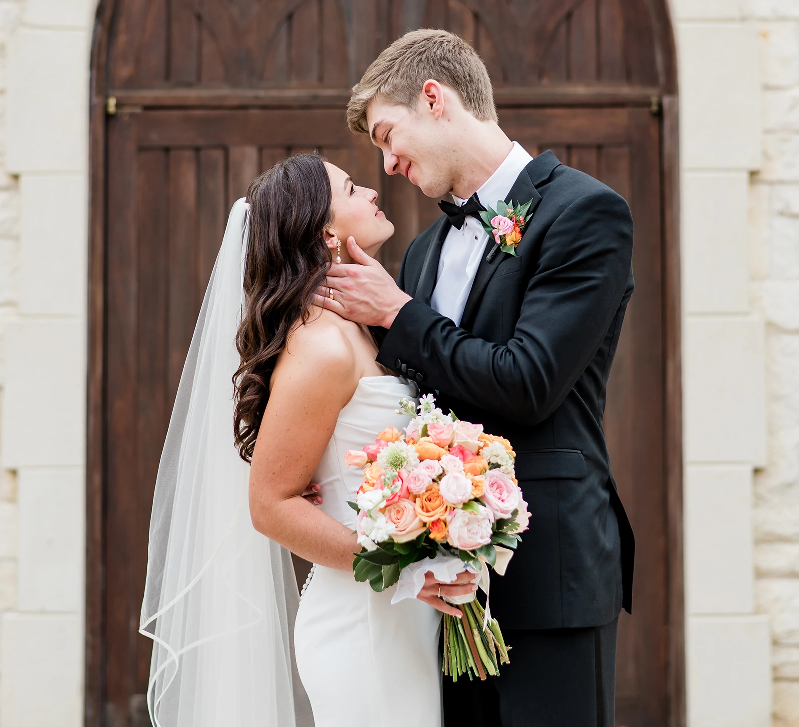 Bride and groom share a romantic moment outside wooden doors, with the bride holding a bouquet of peach and pink roses.