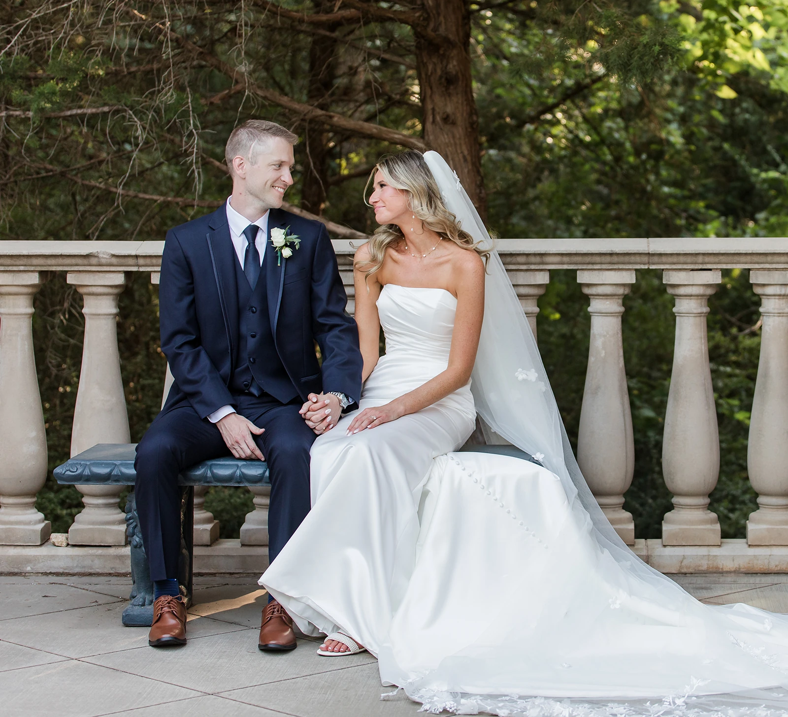 Bride and groom sitting on a stone bench holding hands, smiling at each other during an outdoor wedding portrait
