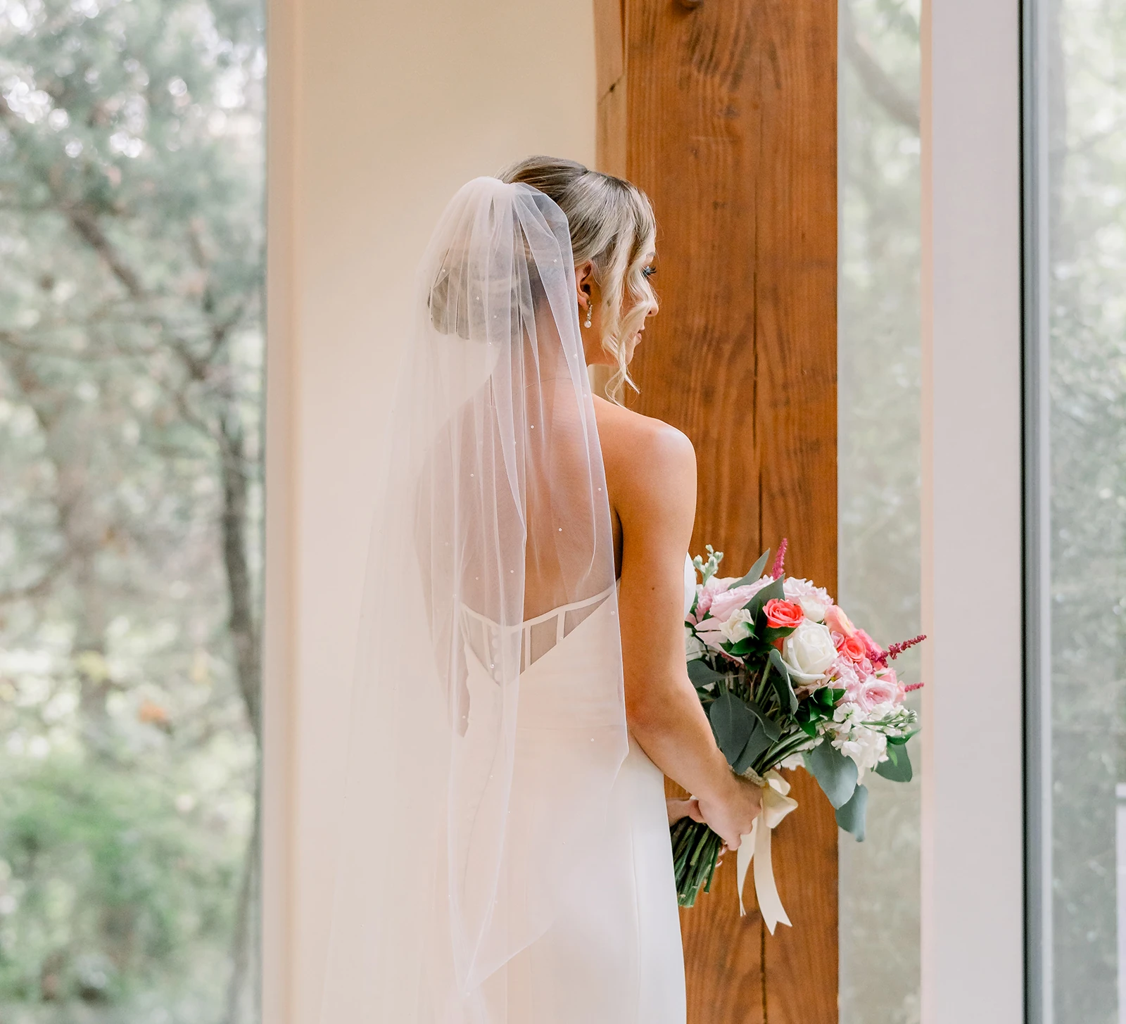 Bride holding a pink and white floral bouquet while standing by a large window with her long veil cascading down her back