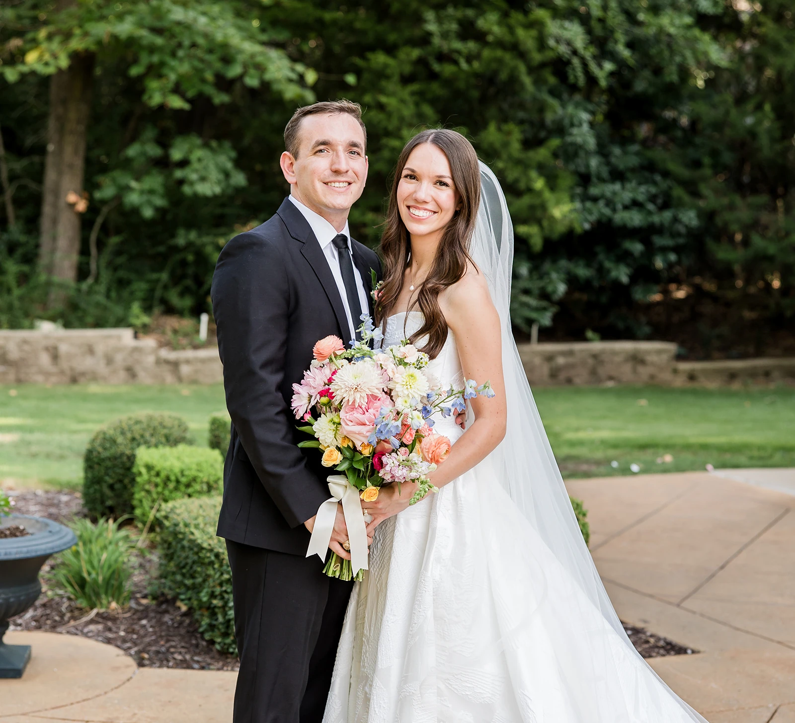 Bride and groom smiling outdoors, with the bride holding a colorful floral bouquet and wearing a long veil