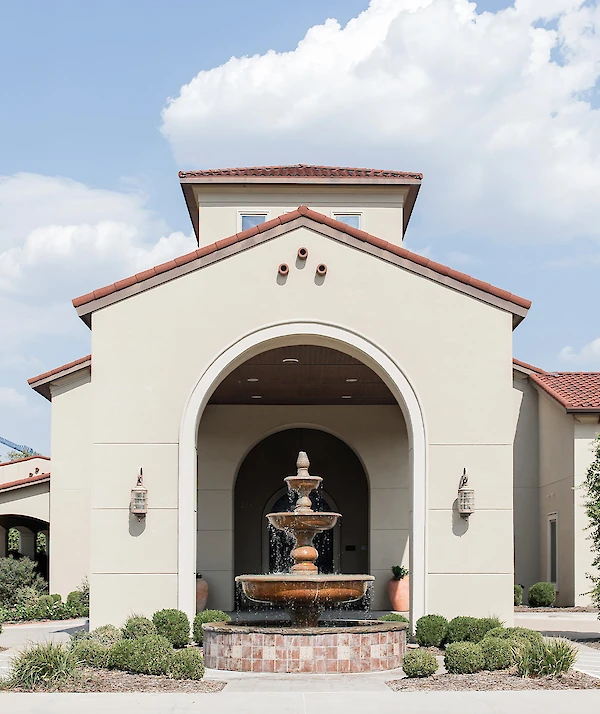 Exterior entrance of a Mediterranean-style event venue with tan stucco walls and a terracotta tile roof, featuring a central arched entryway and a large tiered copper water fountain.